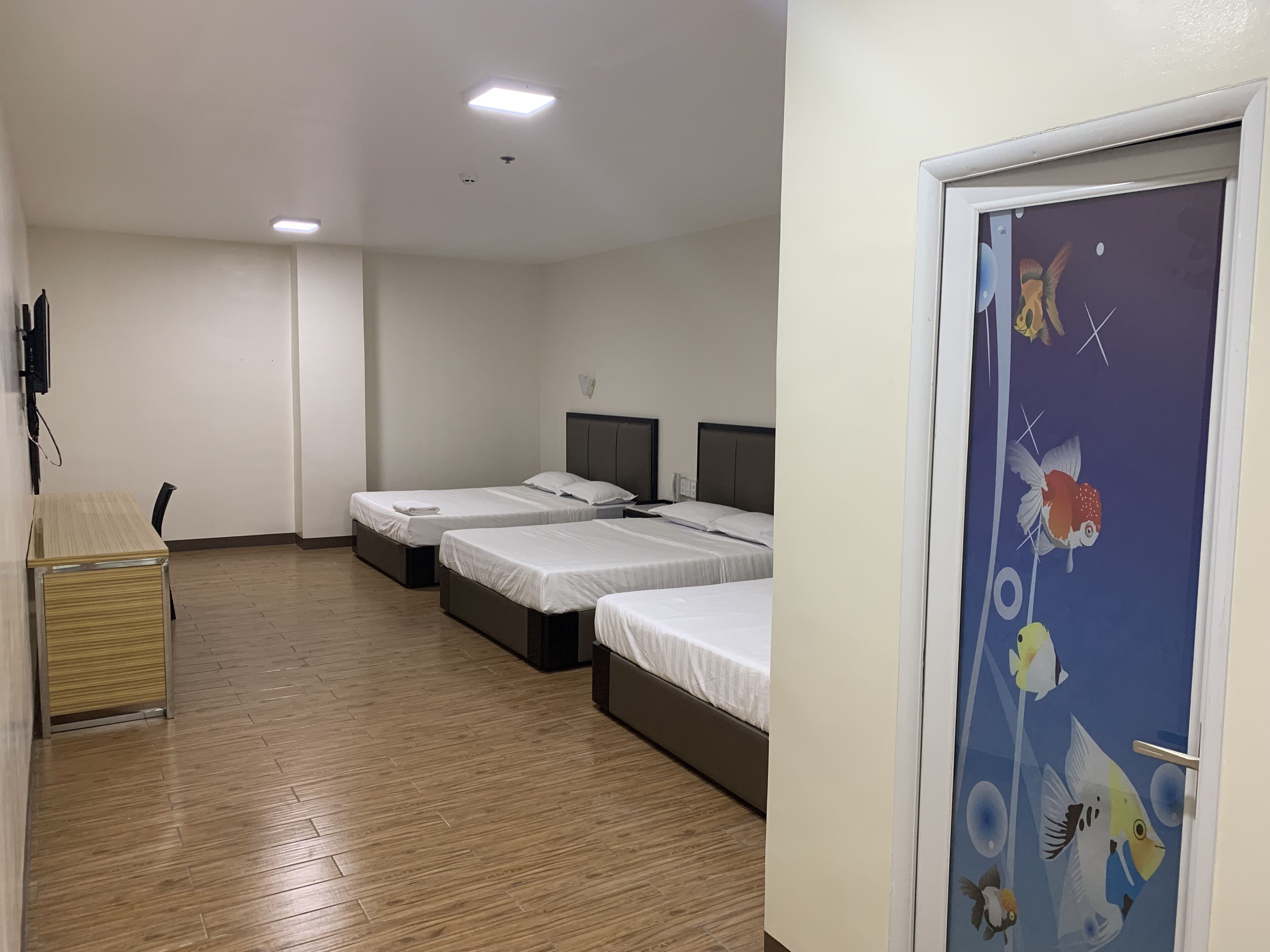 Hotellihuone, jossa on kolme erillistä sänkyä, puulattia, työpöytä tuolilla sekä ovi, joka on koristeltu kalakuvioilla.