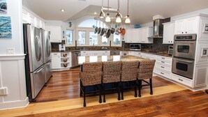 A spacious kitchen with white cabinets, stainless steel appliances, and a central island with wicker chairs. - Oceanside, ELEVATOR, Indoor-Outdoor pool, Sauna, Rec Room, Media Room, BH12, Celtic Tides (Corolla)