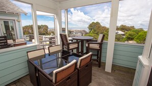 A covered outdoor patio with a glass-top table and wicker chairs, overlooking a residential area with houses and trees. - Oceanside, ELEVATOR, Indoor-Outdoor pool, Sauna, Rec Room, Media Room, BH12, Celtic Tides (Corolla)
