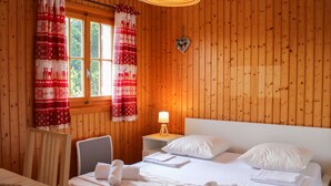 Een gezellige kamer met houten wanden, een bed met witte lakens, een bureau met stoel en een raam met rode gordijnen.
