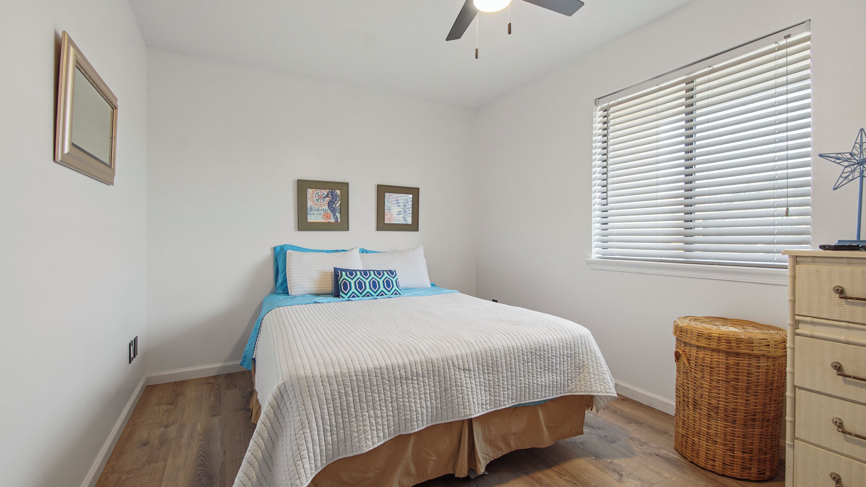 A bedroom with a bed, a ceiling fan, a window with blinds, a wicker basket, and a dresser.