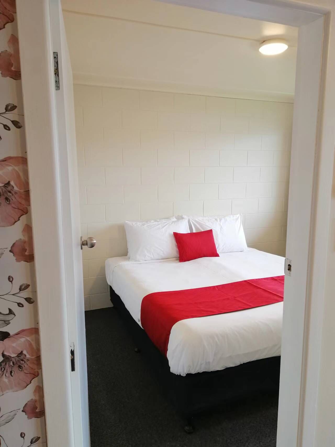 Una habitación de hotel con pared de ladrillo blanco, cama con ropa blanca y almohada roja, y una manta roja.