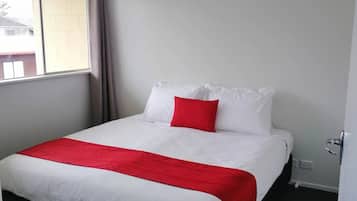 Un lit bien fait, avec une literie blanche et rouge, une fenêtre avec des rideaux et une porte dotée d’une poignée.