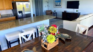 Una cocina moderna con encimeras de granito, armarios de madera y electrodomésticos de acero inoxidable. Se aprecia una zona de comedor con una mesa de madera, sillas blancas y un arreglo floral decorativo. Al fondo, se ve un salón con un sofá blanco y un televisor de pantalla plana.