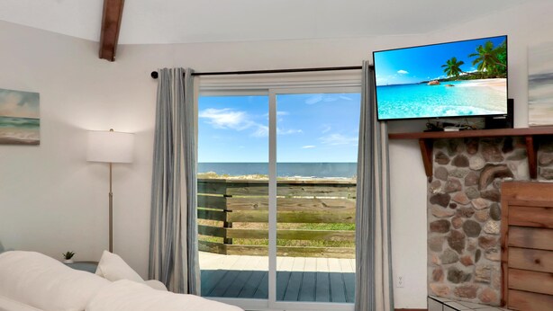 A room with a large sliding glass door leading to a beach view, a flat-screen TV mounted on the wall, a wooden bench, and a stone fireplace. - Amethyst Dunes (Port St. Joe)