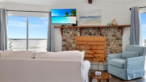 A living room with a stone fireplace, a television, a sofa, a chair, and a view of the beach. - Amethyst Dunes (Port St. Joe)