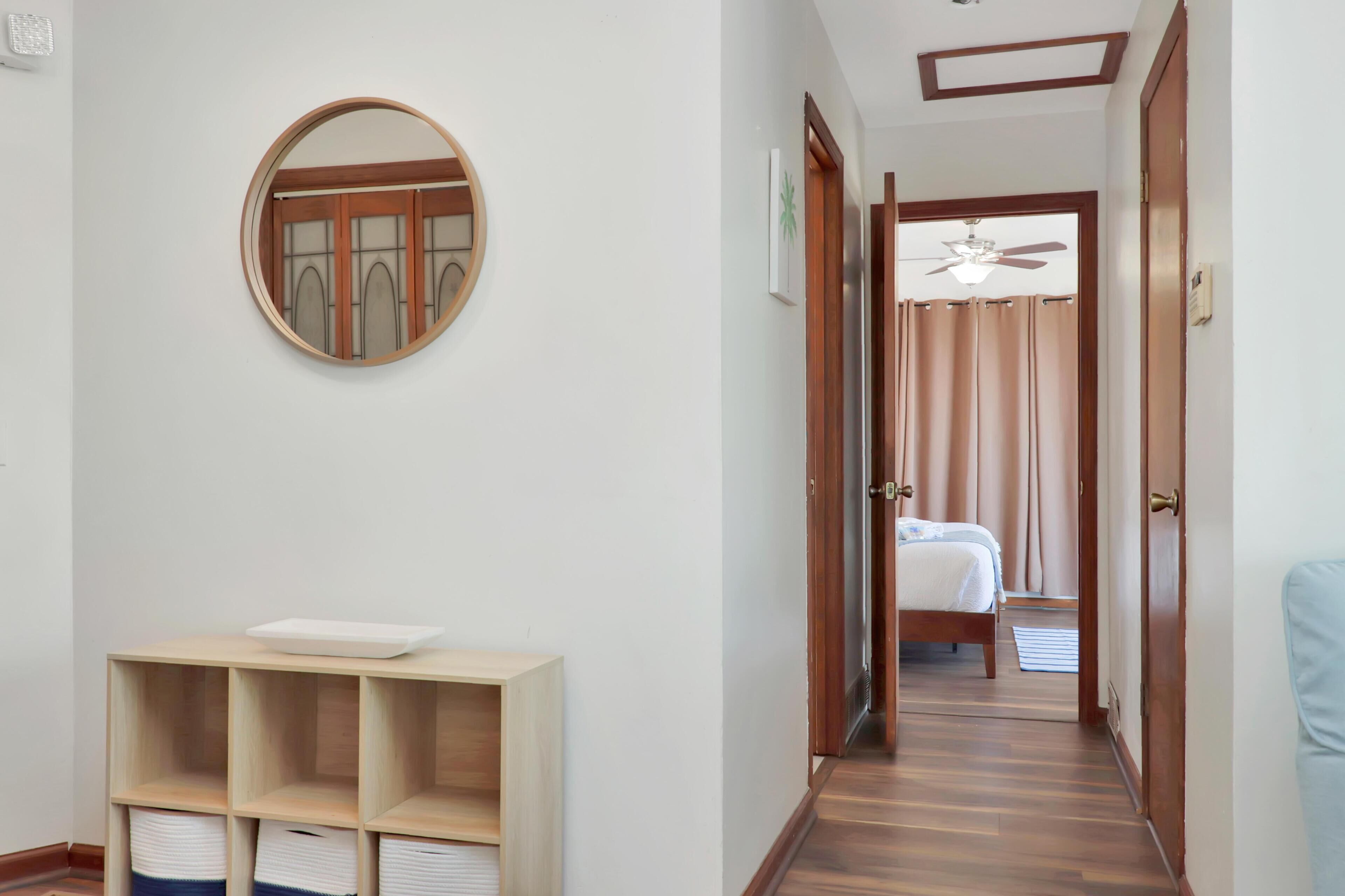 A hallway with a wooden floor, a round mirror on the wall, a bed in the adjacent room, and a ceiling fan.