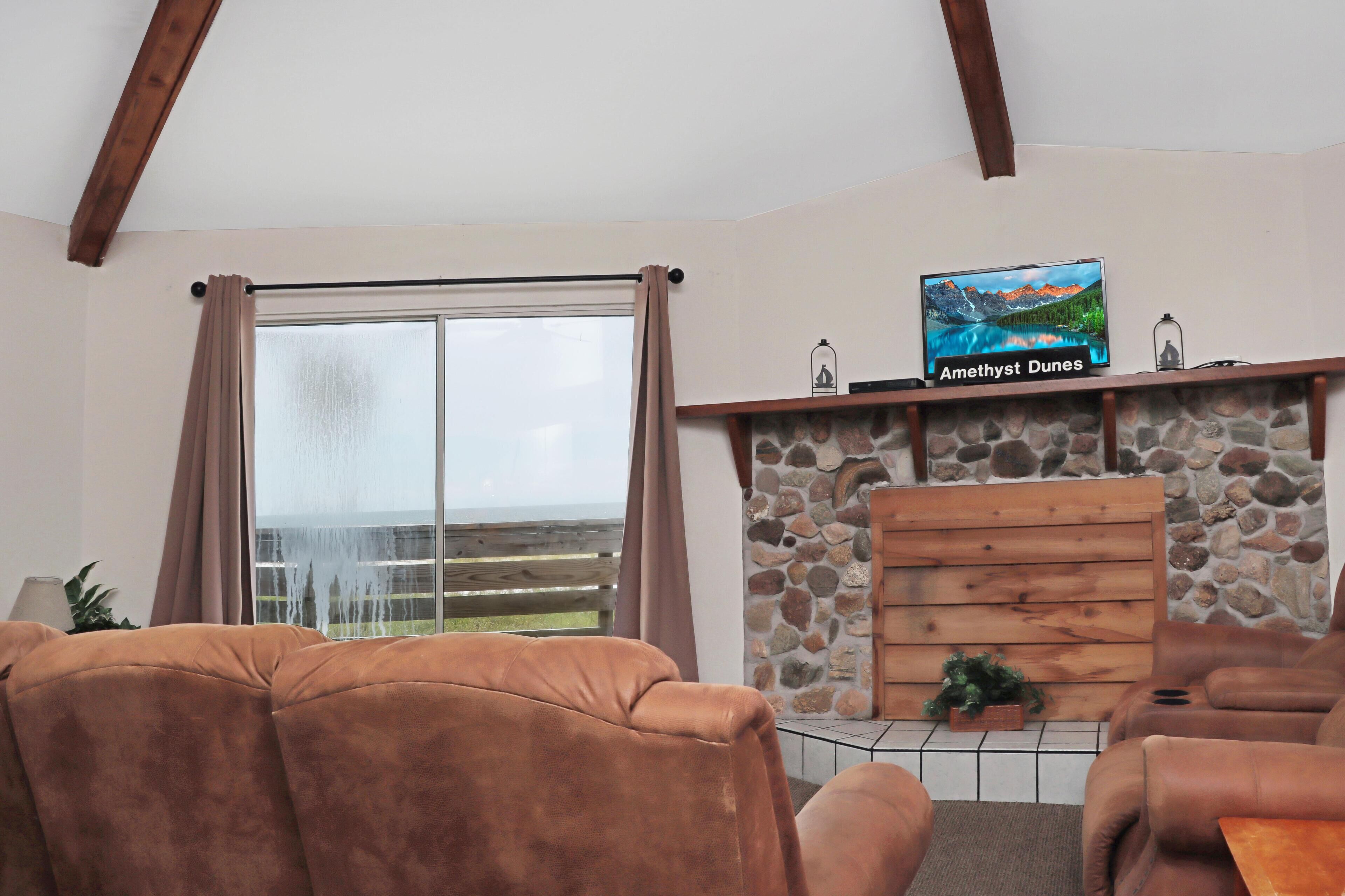 A living room with a stone fireplace, a television mounted above it, and a large window with curtains.