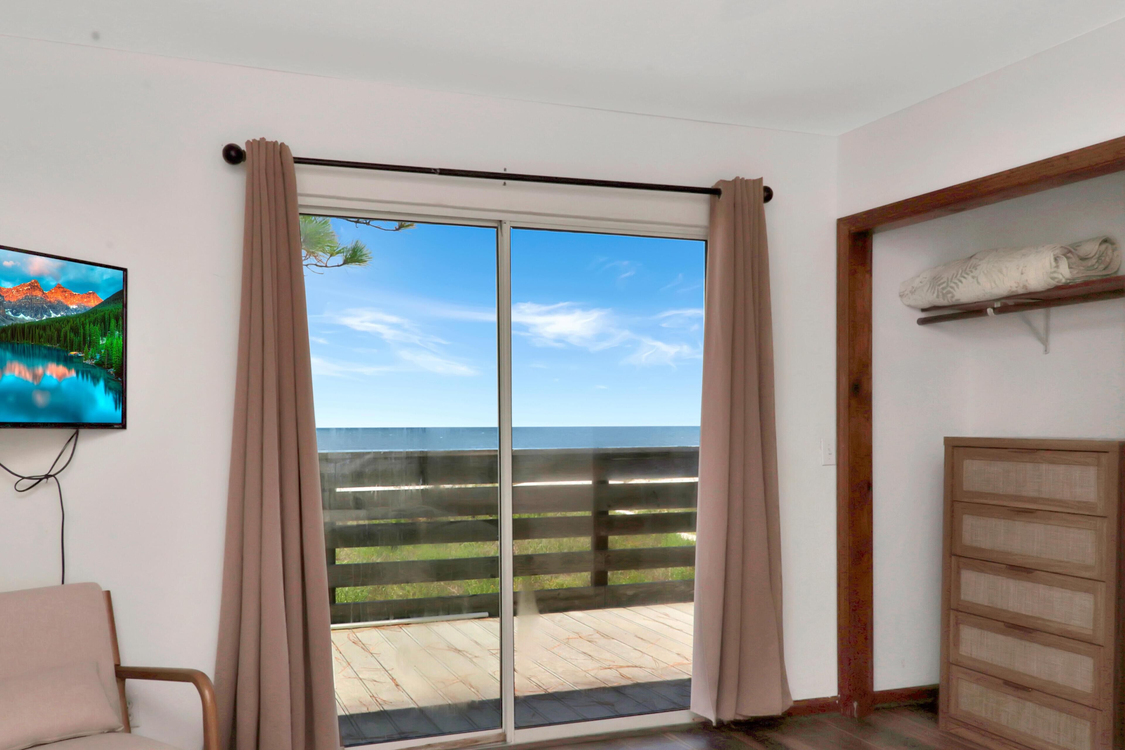 A room with a sliding glass door leading to a balcony with a wooden deck and a view of the ocean.