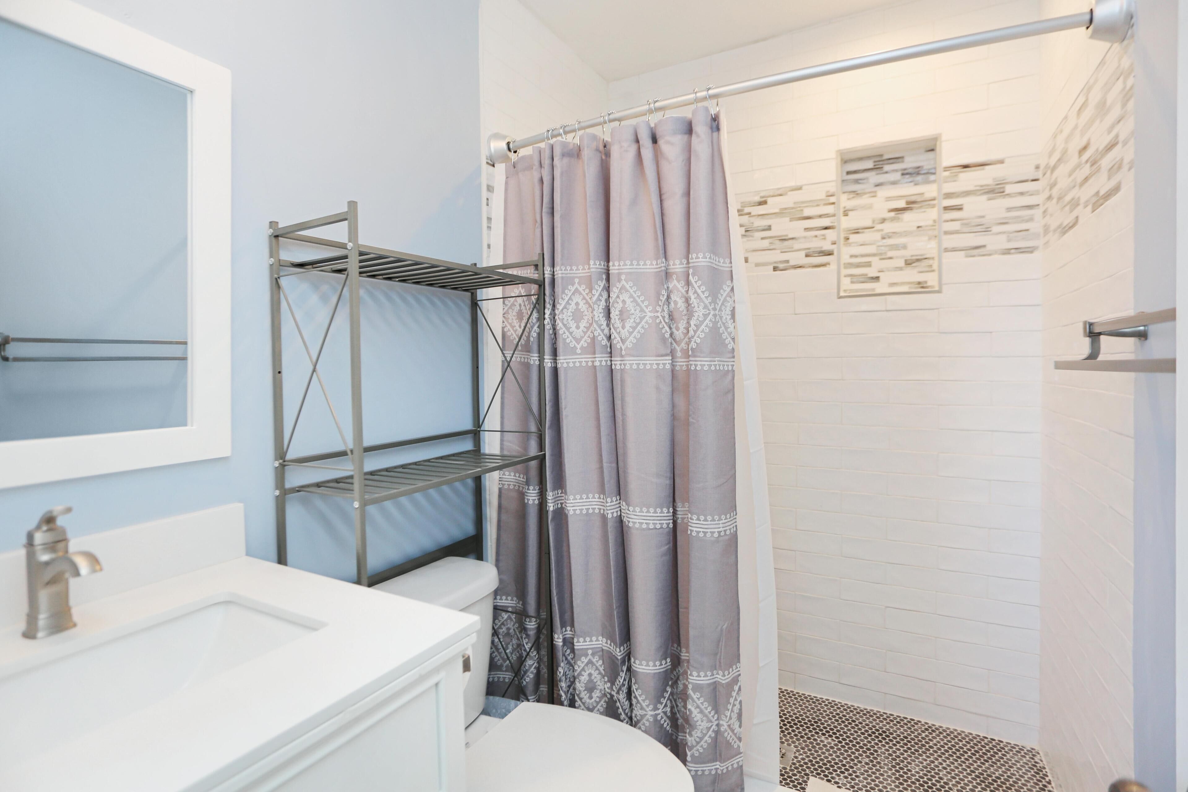 A bathroom with a white sink, a toilet, a shower with a patterned curtain, and a metal shelf unit.