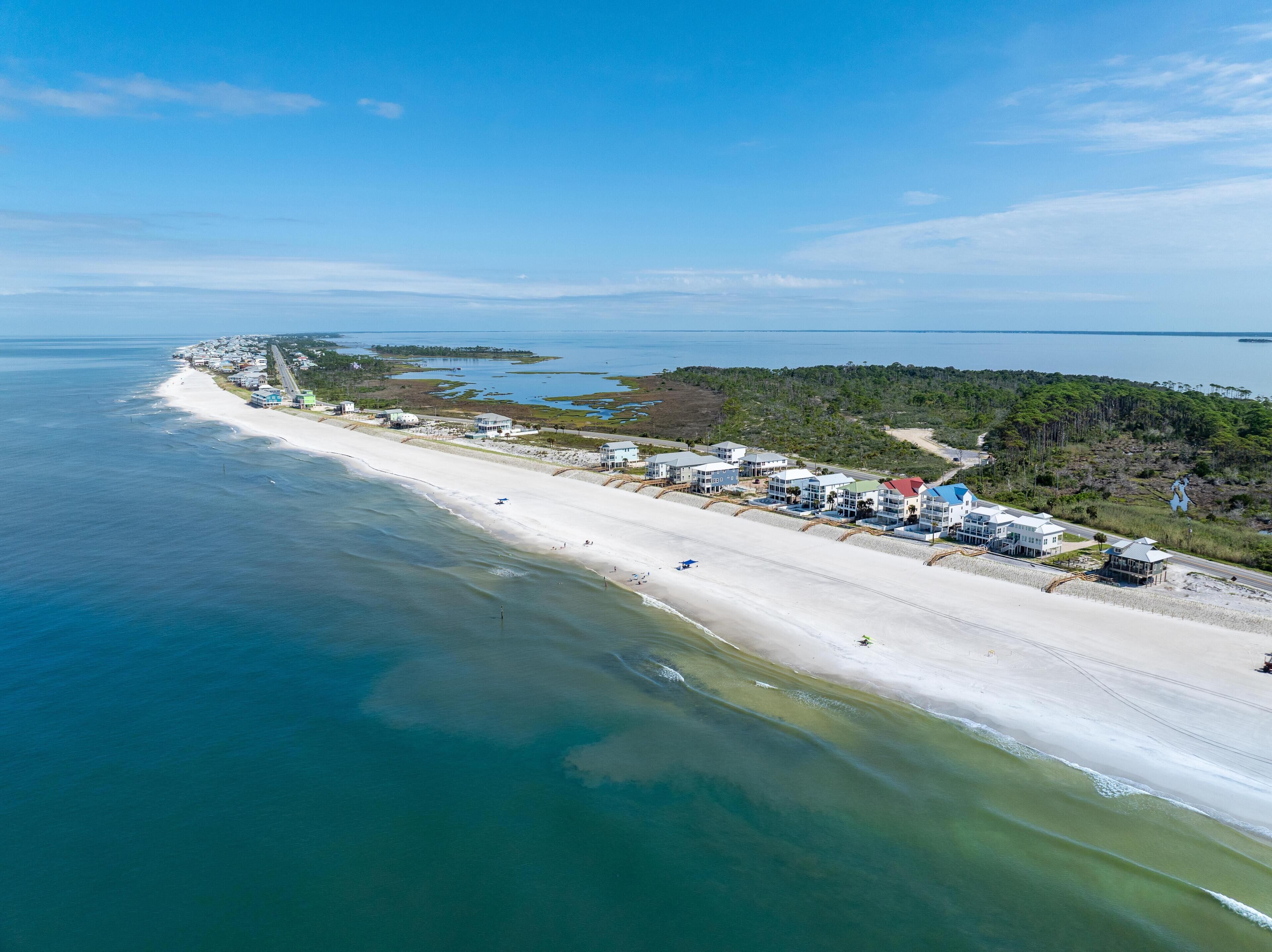 An aerial view of a coastal area with a beach, a row of buildings, and a forested peninsula.