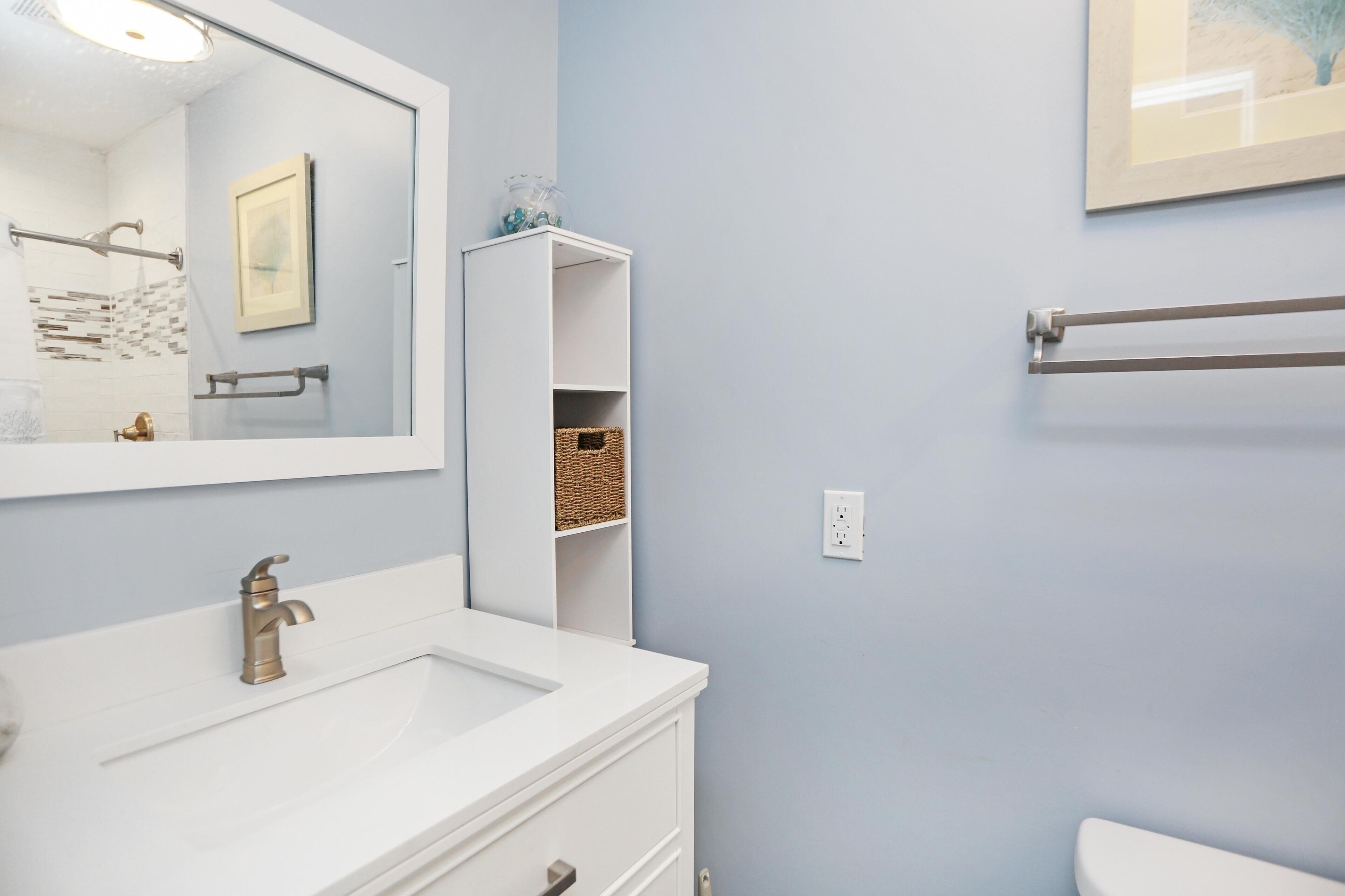 Une salle de bain avec un lavabo blanc, un grand miroir, un porte-serviettes et une étagère avec des paniers.