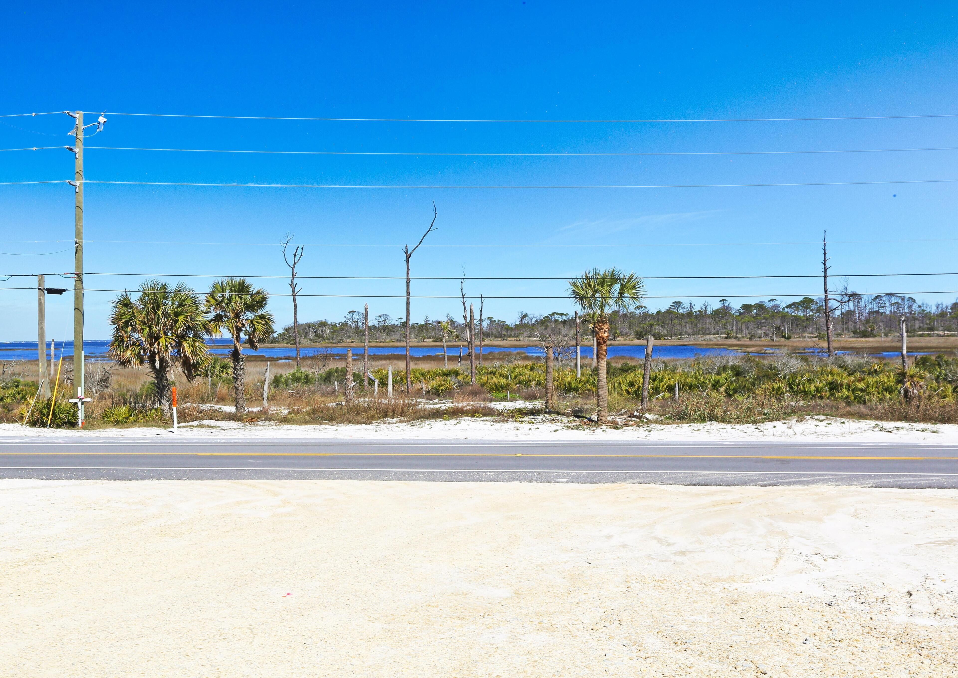 A coastal road with a clear blue sky, a sandy beach, and a body of water in the distance.