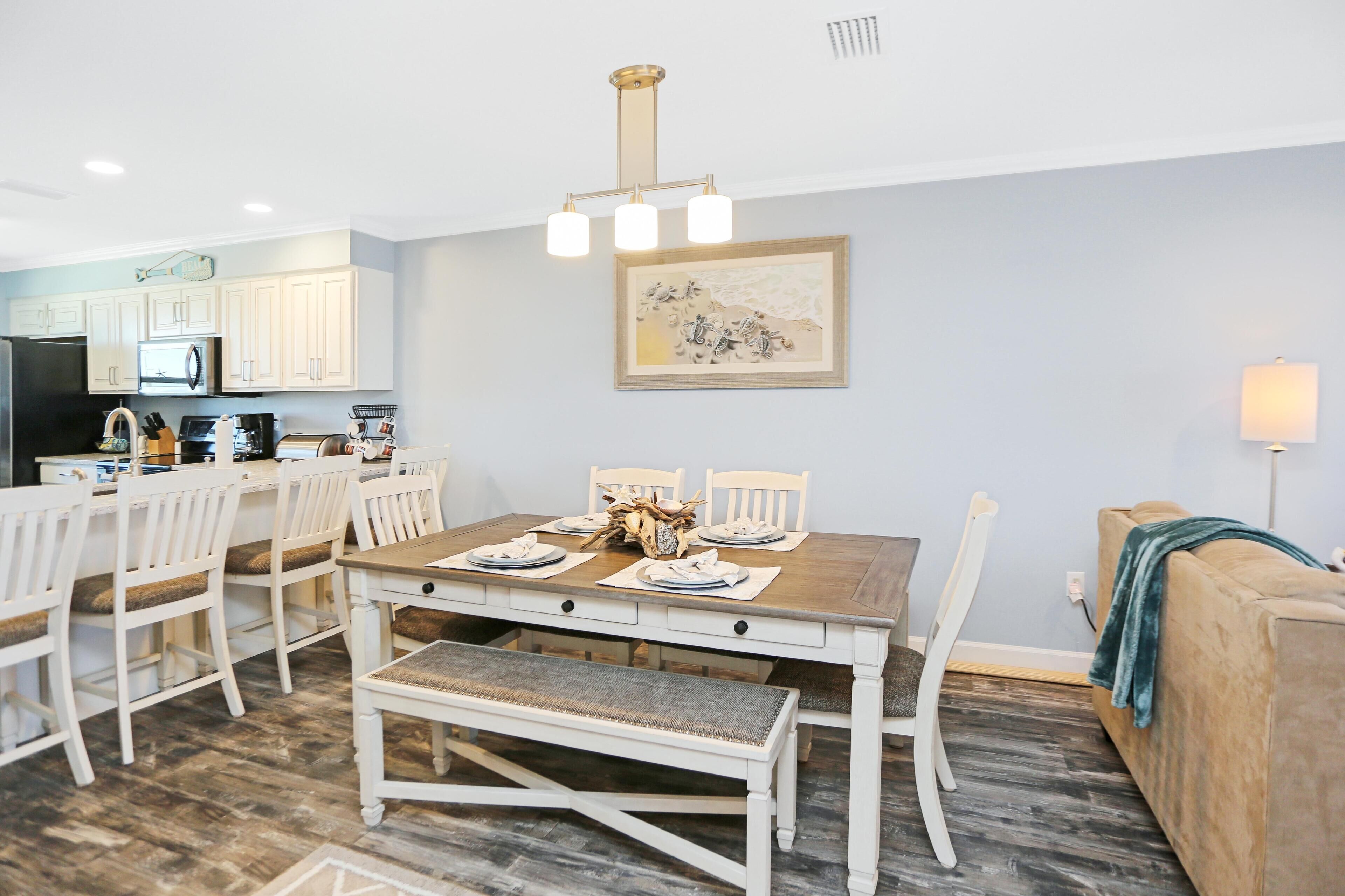 Un comedor con una mesa de madera dispuesta para cuatro personas, rodeada de sillas blancas. Un banco se ubica frente a la mesa. La habitación tiene una pared azul claro y una obra de arte enmarcada sobre la mesa.