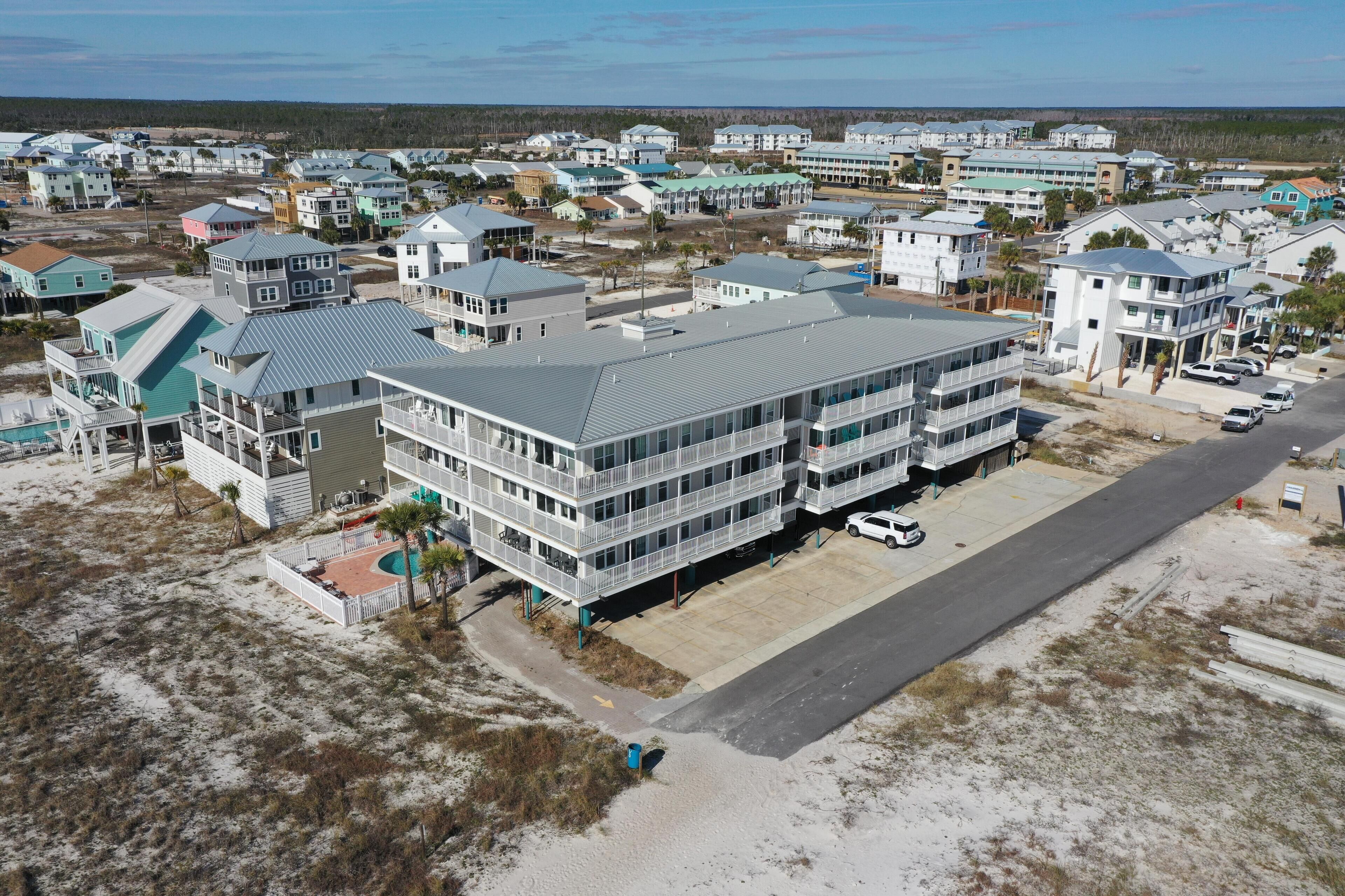 An aerial view of a coastal residential area with multiple two-story houses, a central building with a flat roof, and a clear road.
