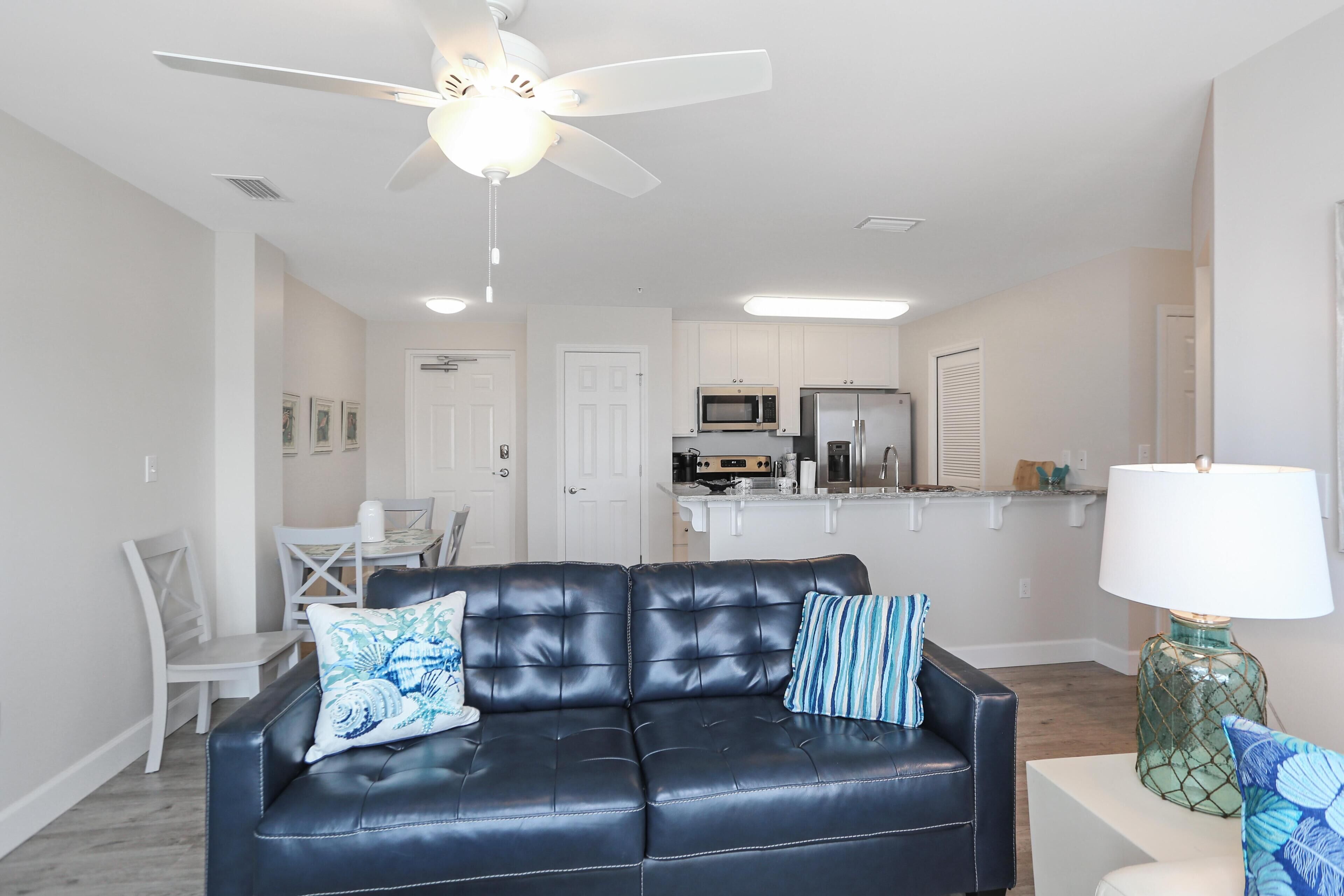 A modern living room with a black leather sofa, a dining table with chairs, a kitchen area with appliances, and a ceiling fan.