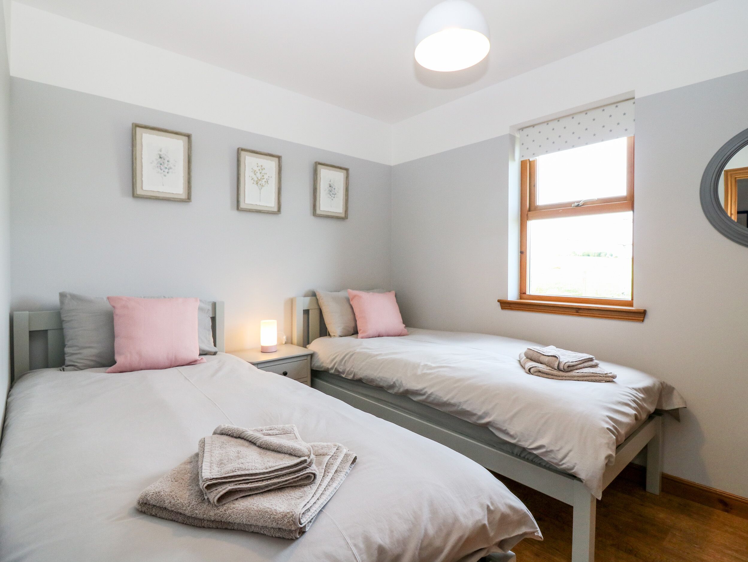 Une chambre avec deux lits, une table de chevet avec une lampe, une fenêtre avec des rideaux et des tableaux encadrés accrochés au mur.