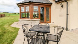 A covered patio area with a table and chairs, a glass-enclosed room, and a view of the surrounding landscape. - Kilnary Cottage (Ellon)