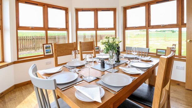 A dining area with a wooden table set for six, large windows with a view of a fenced field, and a pendant light above. - Kilnary Cottage (Ellon)