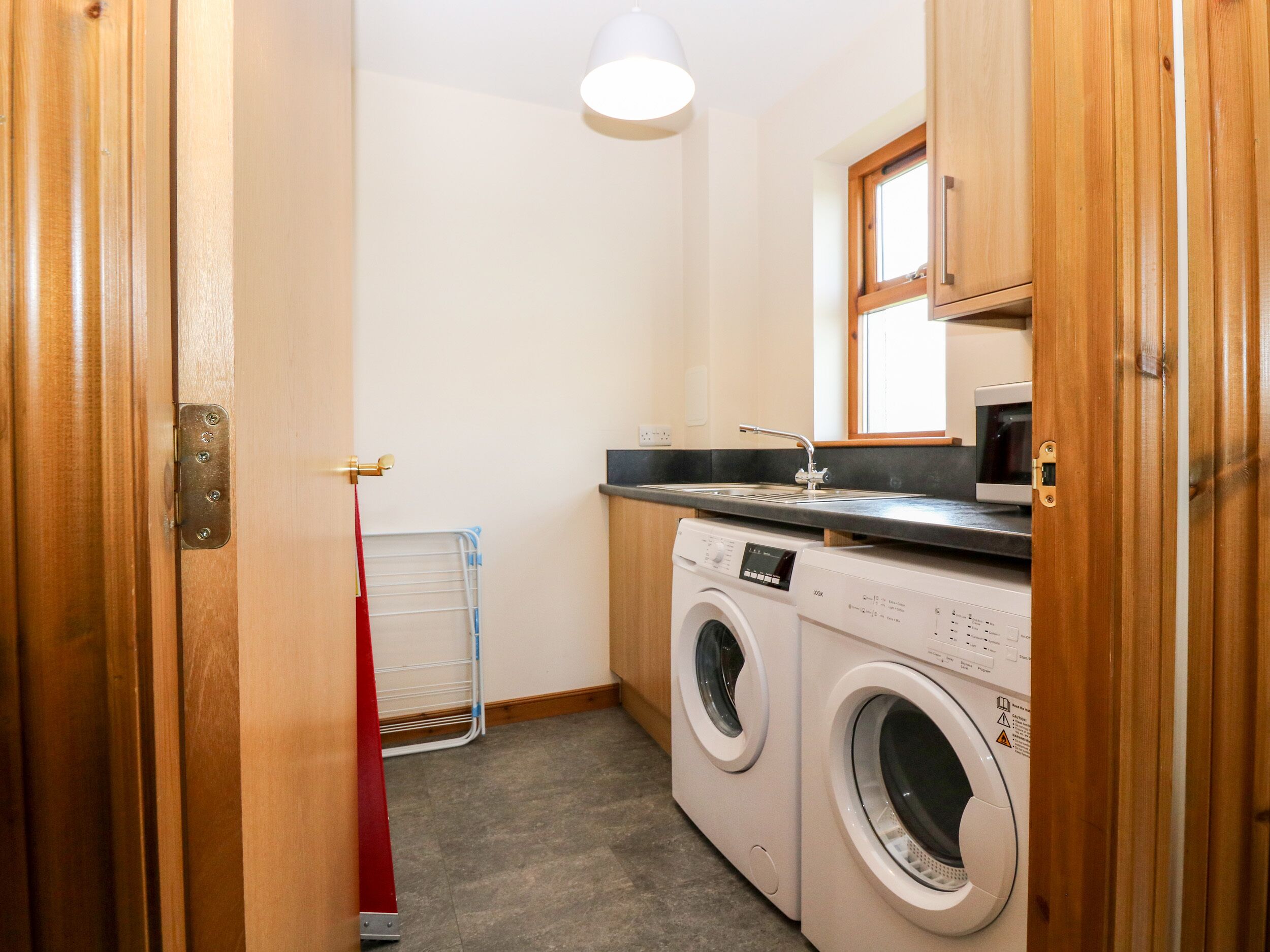 A laundry room with two washing machines, a drying rack, a sink, and a microwave.