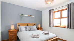 A bedroom with a wooden bed, two bedside tables, a shelf with a plant, and a window with curtains. - Kilnary Cottage (Ellon)