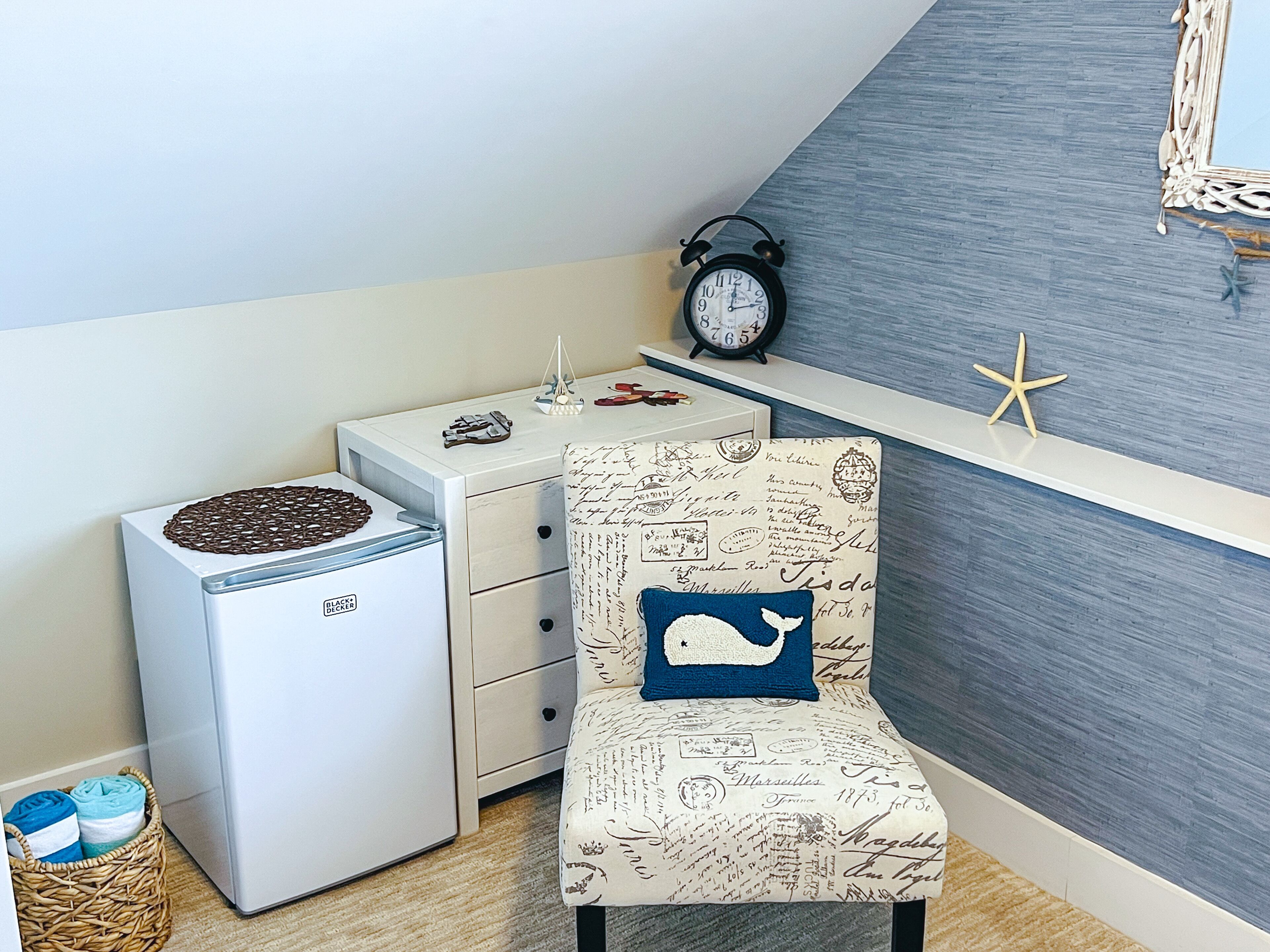 A room with a white chest of drawers, a small heater, a decorative chair with a whale pillow, a wall clock, and a starfish decoration.