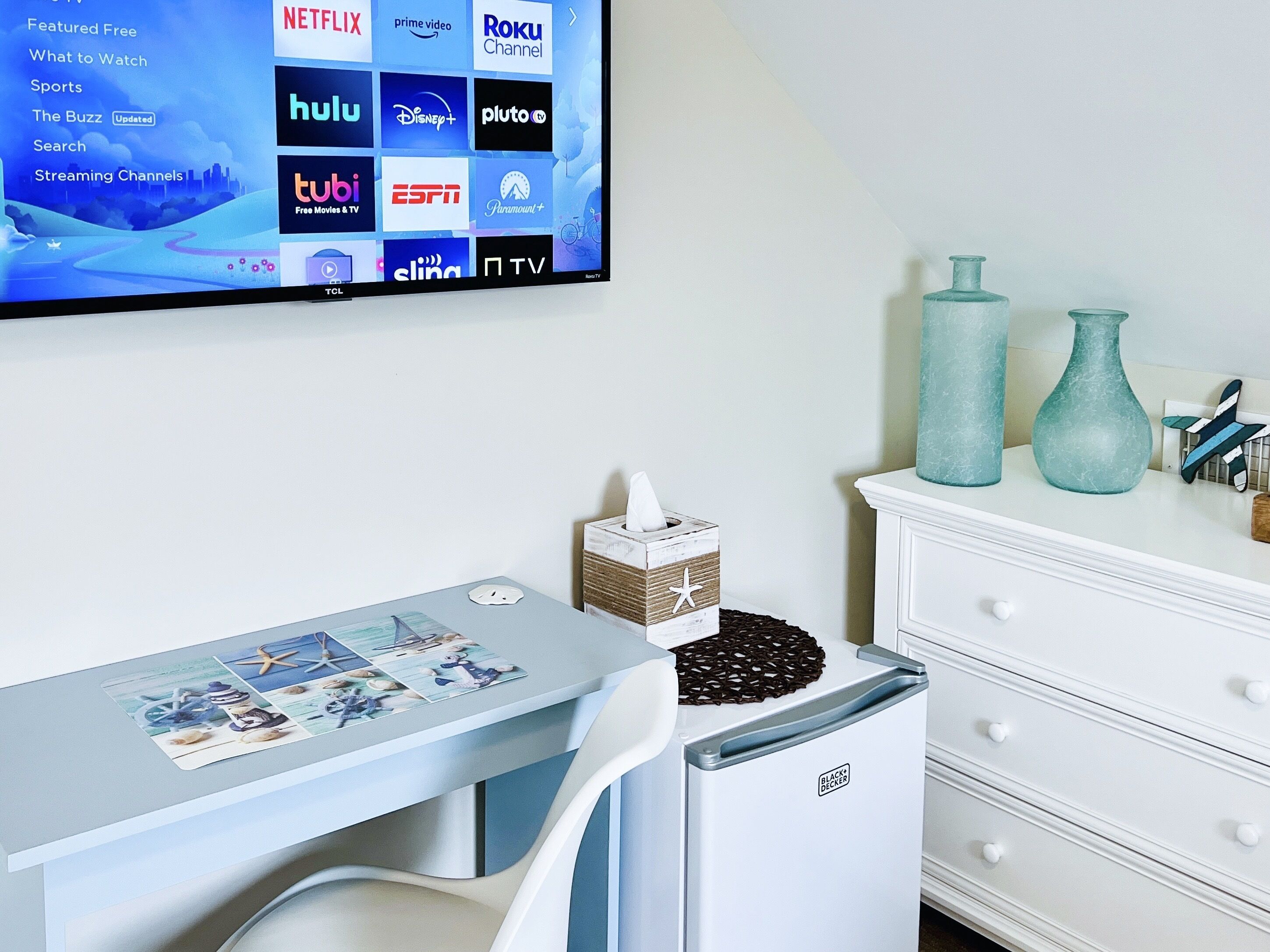 Una habitación moderna con una televisión de pantalla plana mostrando servicios de transmisión, un escritorio con posavasos, una caja de pañuelos, jarrones decorativos y un aparador blanco.