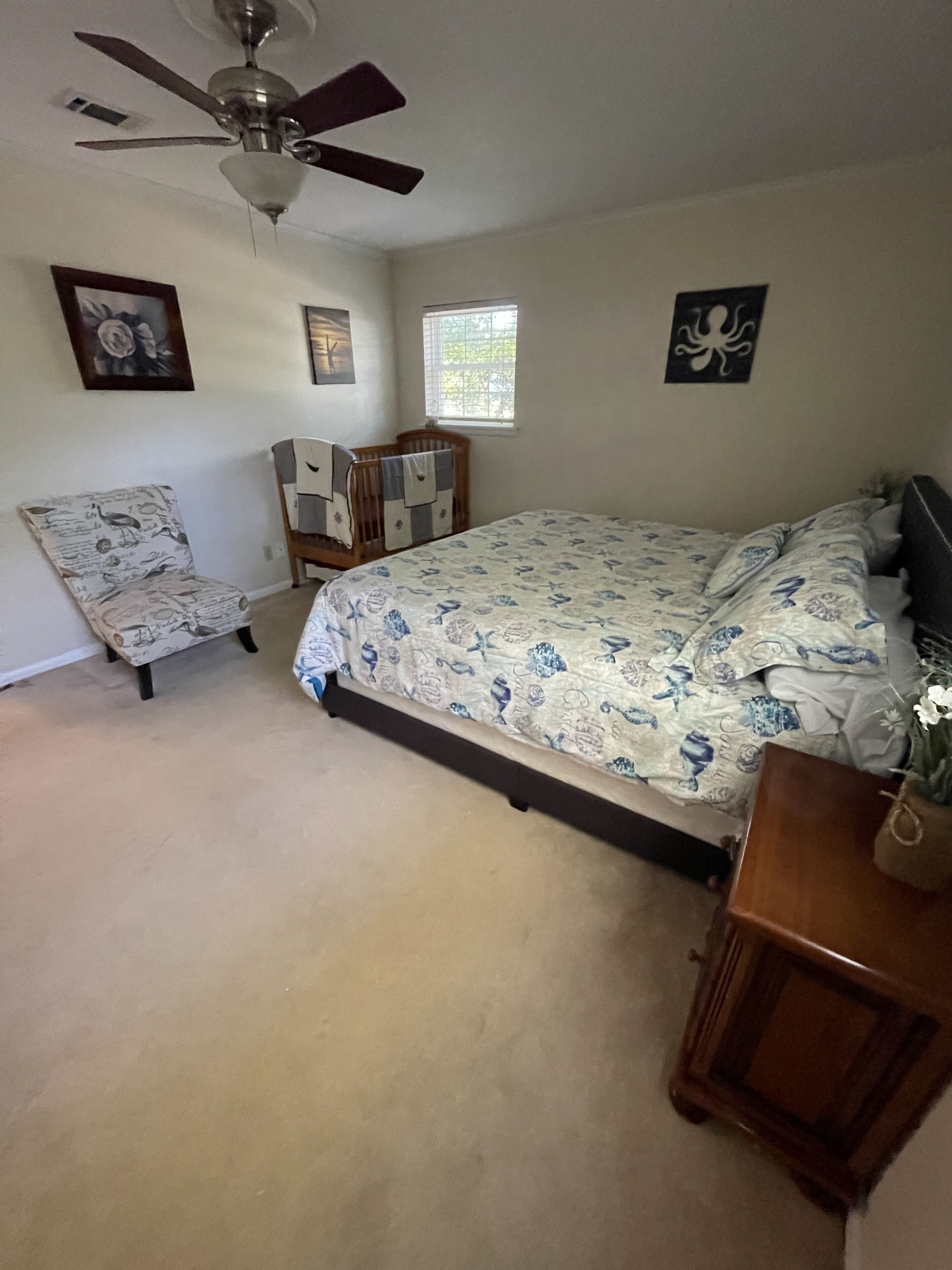 A bedroom with a bed, a chair, a crib, a ceiling fan, and a window.