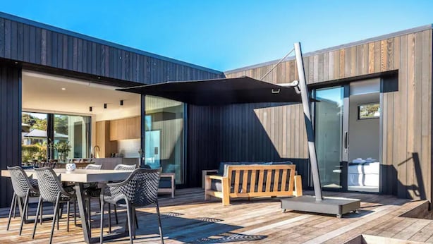 A modern outdoor patio with a wooden deck, a dining table with chairs, a bench, and a glass door leading to a covered area. - 2 min walk to the glorious white sands of Onetangi beach (Waiheke Island)