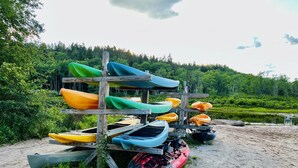 A wooden rack holding various kayaks and a red canoe on a sandy shore with a forested background.