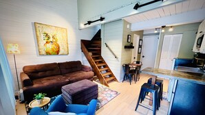 A cozy living room with a brown sofa, a blue armchair, and a wooden staircase leading to a kitchen area.