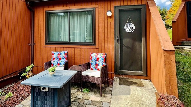 A wooden house with a tiled roof, a front door with a 'hello' sign, a small porch with a table and two chairs, and a garden area with a potted plant.