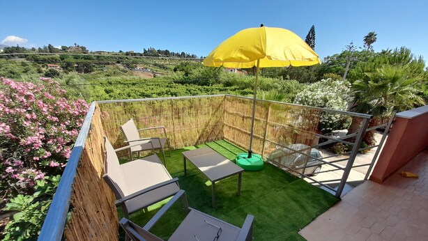 Une terrasse avec une table et des chaises, un parasol jaune et une vue sur un jardin où poussent des fleurs roses.