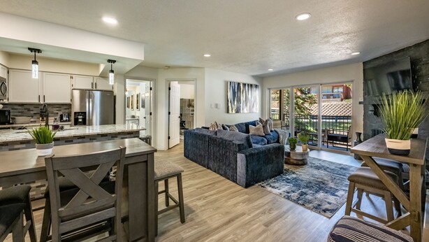 A modern kitchen with stainless steel appliances, a dining area with a wooden table and chairs, and a living room with a blue sofa and a large window. - Total Renovation 300' from Chili Express (Angel Fire)