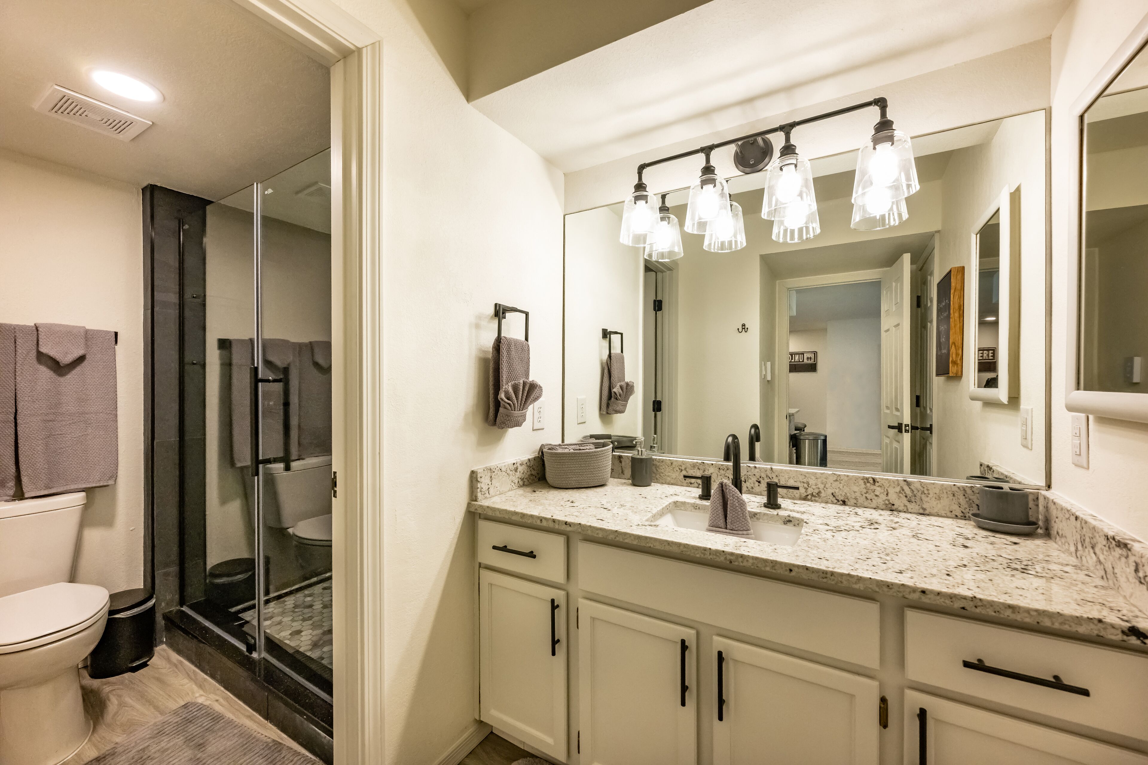 A modern bathroom with a glass shower enclosure, a white sink with a marble countertop, and a large mirror with overhead lighting.