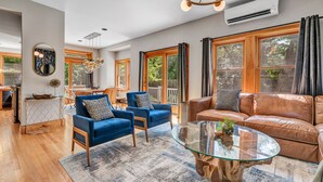 A spacious living room with a glass coffee table, blue armchairs, a brown leather sofa, and large windows with curtains.