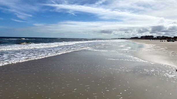 Une plage avec des vagues, un ciel dégagé et des bâtiments visibles au loin.