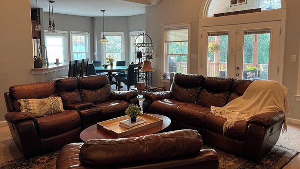 A spacious living room with a brown leather sectional sofa, a coffee table, and a dining area in the background. - Island home on the water with pool.  Bring your boat or jet skis ! Golf near by (Currituck)