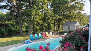 A backyard with a swimming pool, surrounded by trees and colorful Adirondack chairs. - Island home on the water with pool.  Bring your boat or jet skis ! Golf near by (Currituck)
