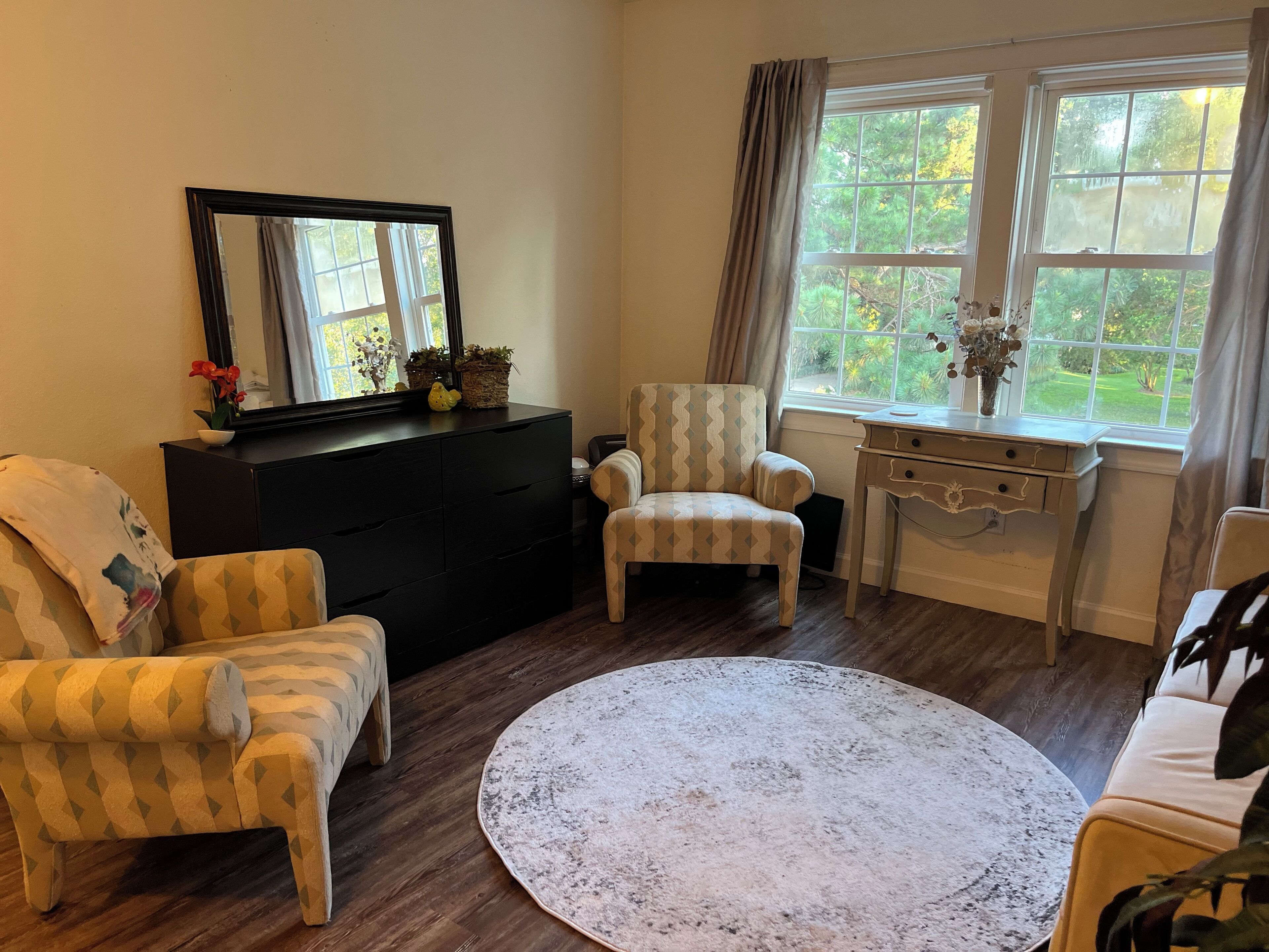 Ein Wohnzimmer mit einem runden Teppich, zwei Sesseln, einem Sofa, einer Kommode, einem Schreibtisch und einem großen Fenster mit Vorhängen.