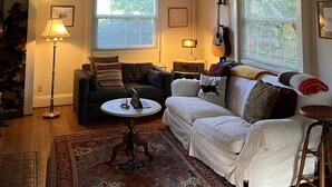 A cozy living room with a sofa, a small round table, and a lamp. There is a guitar leaning against the wall and a window with blinds. - ♥️ of UofO pristine residence for the World Championships (Eugene)