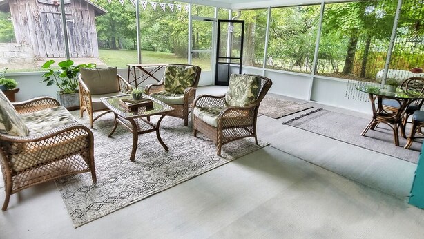 Une terrasse couverte, aménagée avec des meubles en osier, une table en verre et offrant une vue sur une structure en bois et de la verdure.