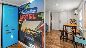 A room with a blue refrigerator, a neon sign for a motel, wooden flooring, a table with chairs, and a bookshelf. - Downtown Big Sky Bungalow w/AC, Fully Fenced Yard, W/D, 2 Smart TVs, Speedy WiFi (Flagstaff)