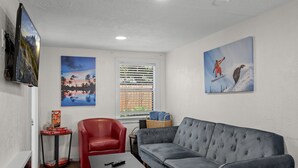 A living room with a grey sofa, a red chair, a small table, and a TV mounted on the wall. There are two paintings on the walls and a window with blinds. - Downtown Big Sky Bungalow w/AC, Fully Fenced Yard, W/D, 2 Smart TVs, Speedy WiFi (Flagstaff)