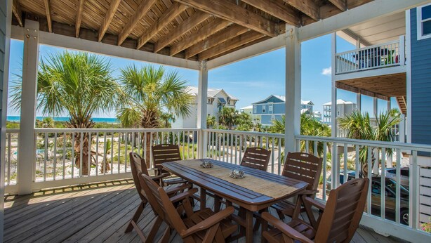 A covered outdoor deck with a wooden table and chairs, overlooking a beach with palm trees and houses. - Veranda Views (Port St. Joe)