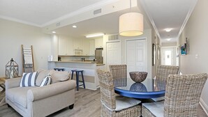 Een moderne woonkamer met een bank, rieten stoelen, een glazen tafel, en een keuken op de achtergrond.