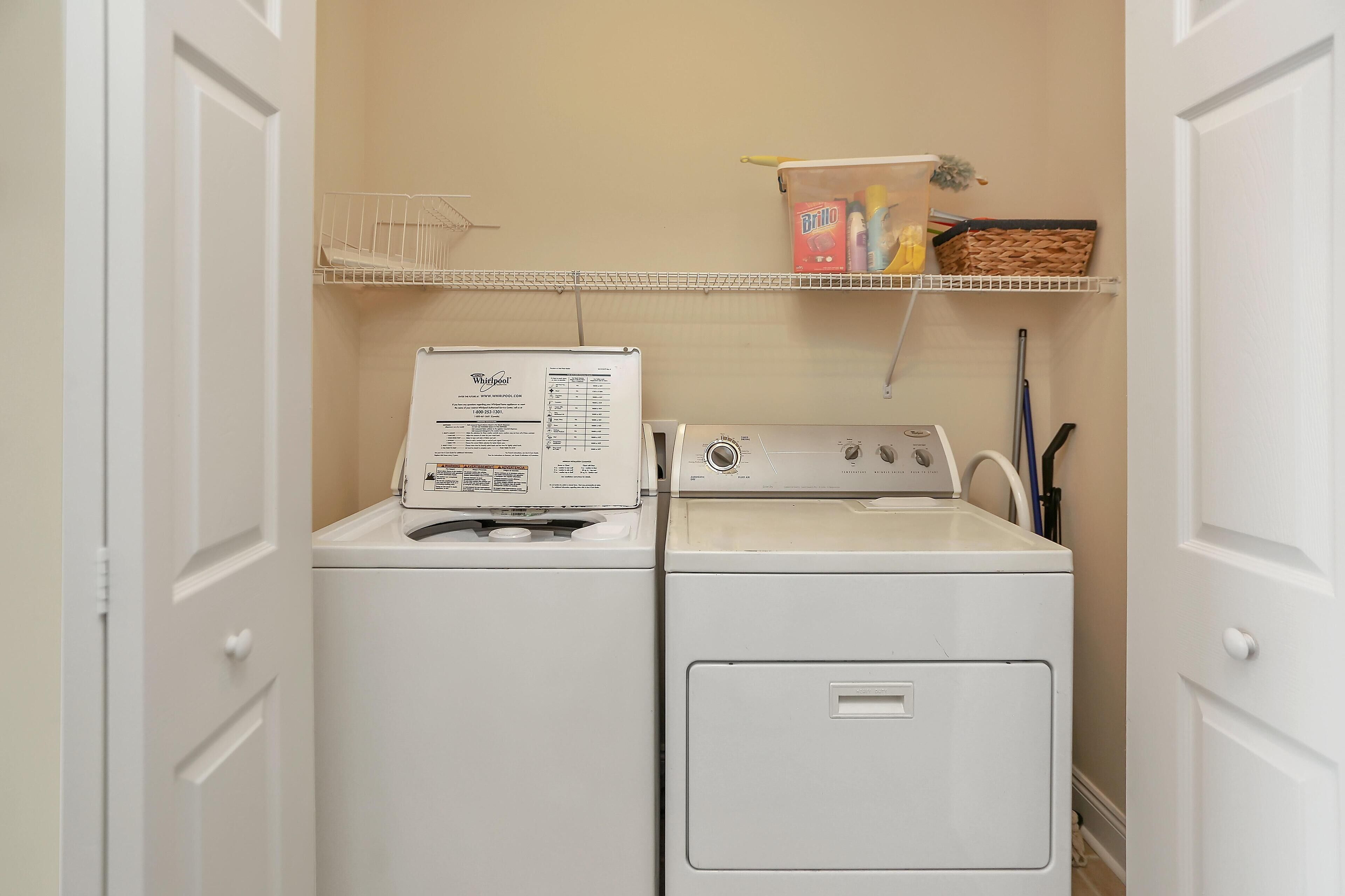 A laundry room with a front-loading washer and dryer, a shelf above with cleaning supplies, and a door on the left.