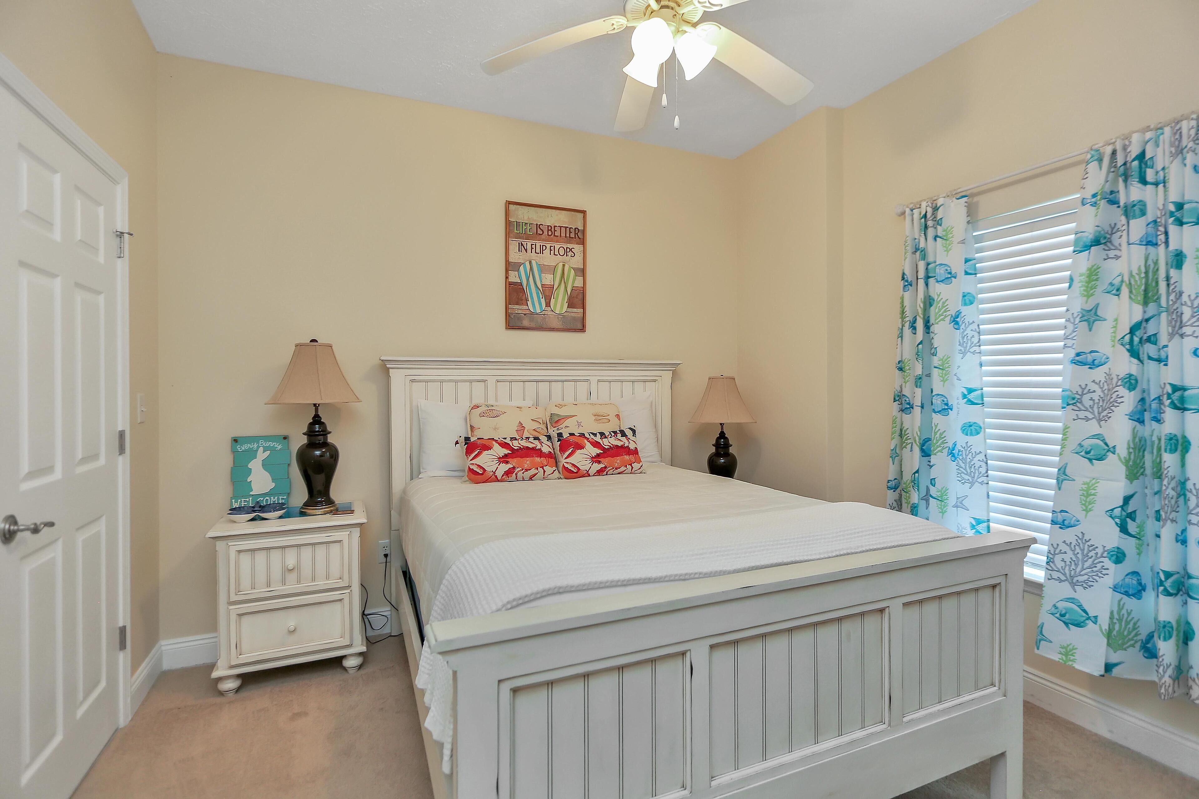 A bedroom with a white bed, two bedside lamps, a small nightstand, a ceiling fan, a window with curtains, and a wall poster.
