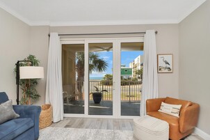 Una sala de estar con un sofá azul, un sillón naranja, una lámpara de pie y vistas a un balcón con una palmera y un edificio verde al fondo.