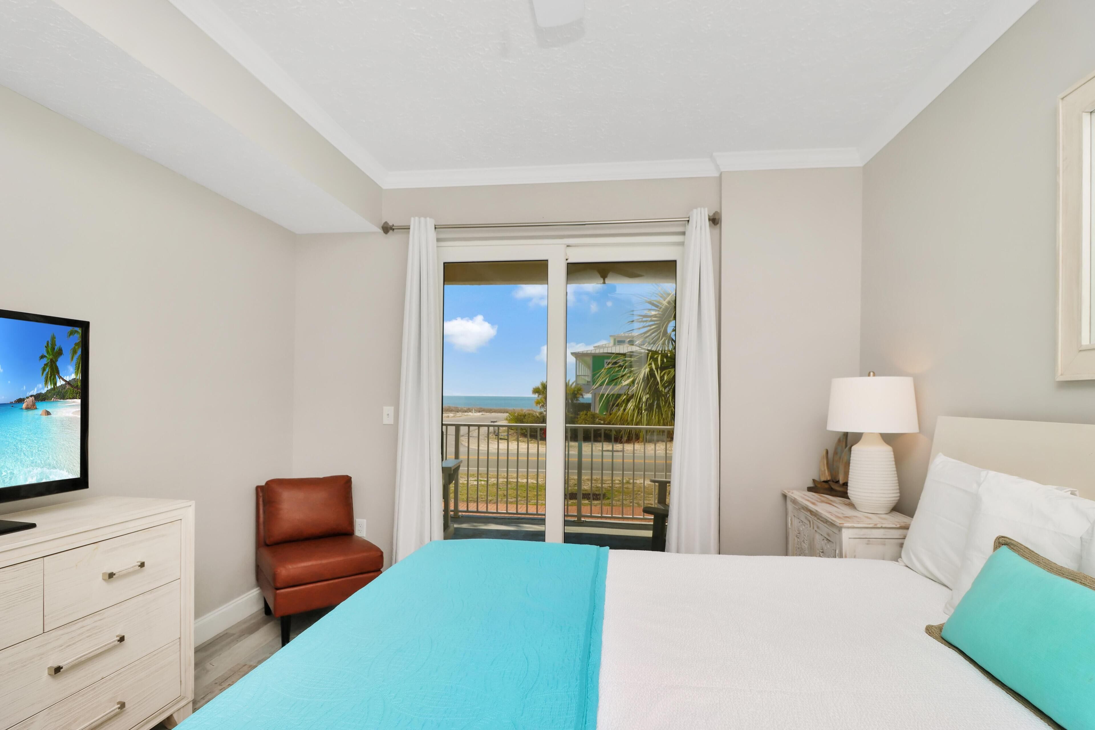 A hotel room with a bed, a TV, a chair, a nightstand, and a view of the beach through a sliding door.
